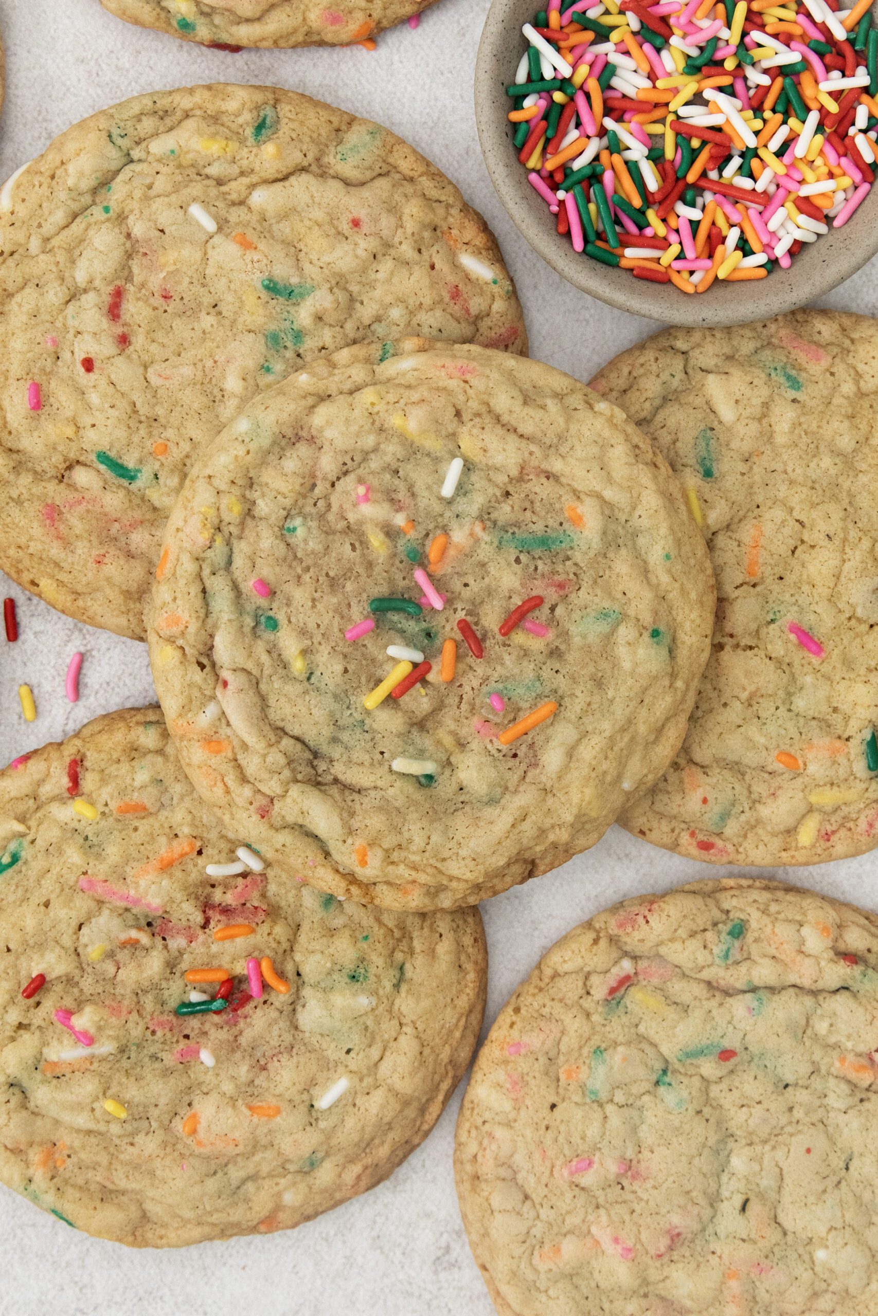 Large sugar cookies with colorful rainbow sprinkles scattered on top, arranged on a light surface. A small bowl filled with extra sprinkles sits beside the cookies.
