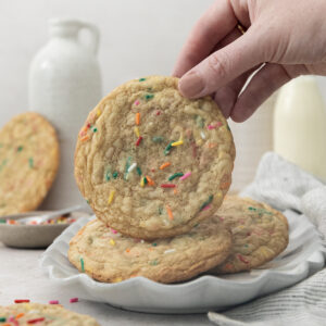 A hand holds a large sugar cookie with colorful sprinkles above a plate stacked with similar cookies; a jug and glass of milk and more cookies are in the background.