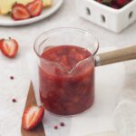 A glass saucepan filled with strawberry sauce sits on a wooden board, surrounded by fresh strawberries, a slice of cake on a plate, and a white container brimming with ripe strawberries in the background.