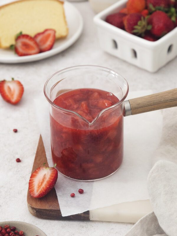 A glass saucepan filled with strawberry sauce sits on a wooden board, surrounded by fresh strawberries, a slice of cake on a plate, and a white container brimming with ripe strawberries in the background.