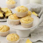 Six pineapple muffins in white paper liners are arranged on a white cake stand, with extra muffins, pineapple slices, and a glass of milk in the background. A striped cloth adds a cozy touch to this delicious muffins display.