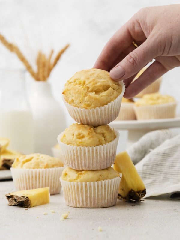 A hand places a muffin on top of a stack of three pineapple muffins, surrounded by pineapple slices, with more muffins and a glass of milk in the background—perfect for muffin lovers.