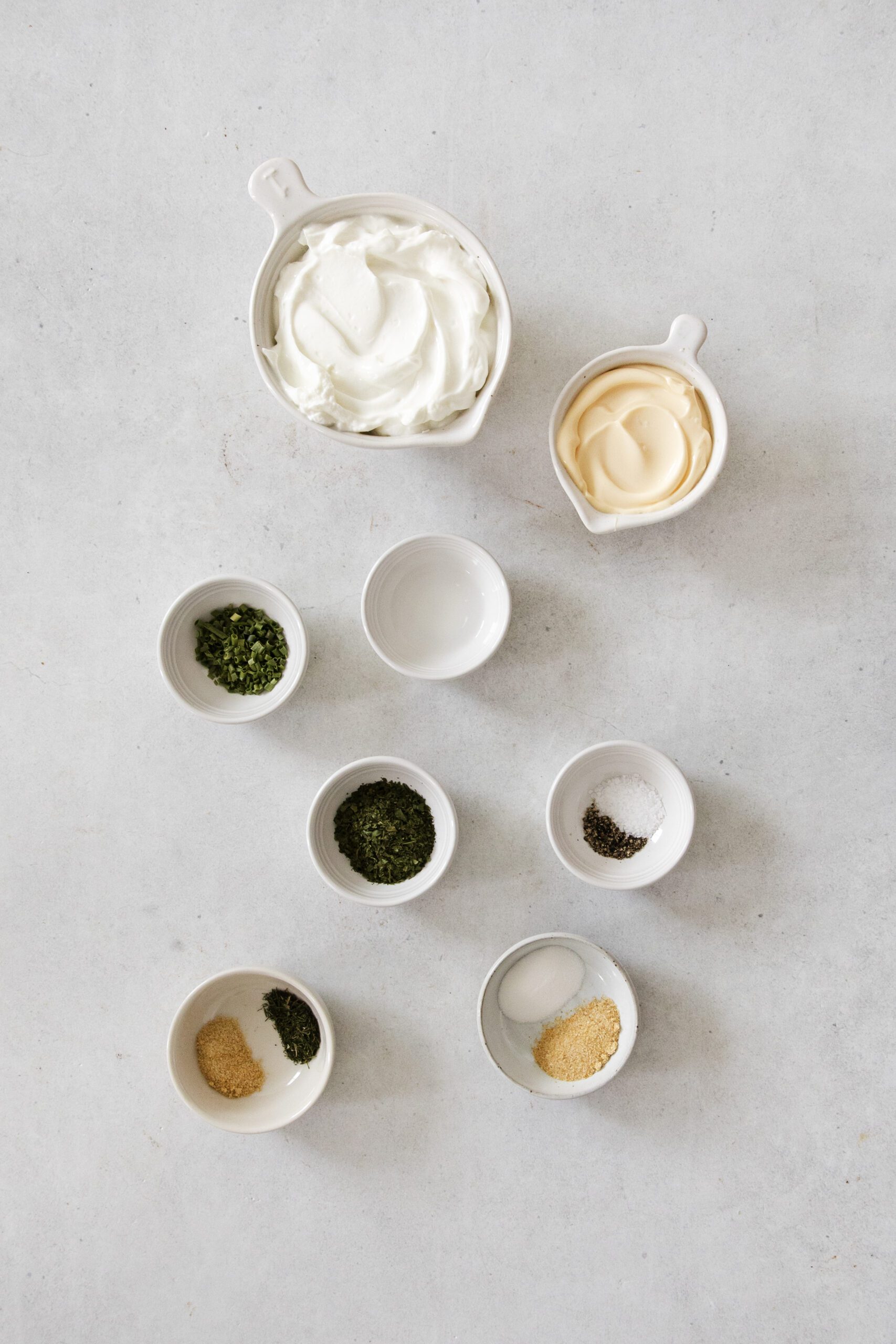An assortment of small white bowls on a light surface, each containing ingredients for a creamy veggie dip—sour cream, mayonnaise, dried herbs, spices, and a clear liquid—arranged neatly as if prepping homemade ranch dressing.