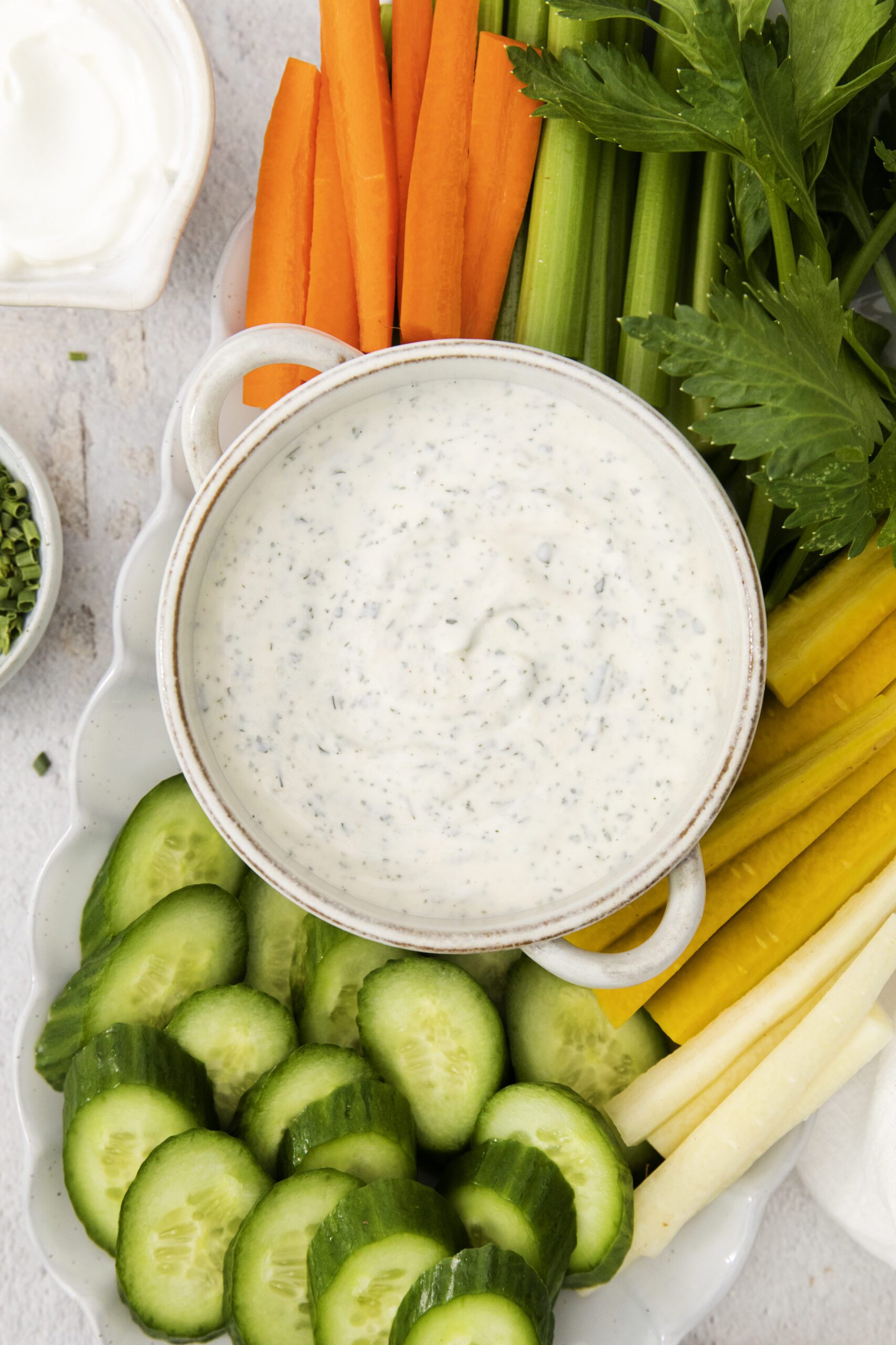 A bowl of creamy ranch dip made with tangy ranch dressing is surrounded by fresh sliced cucumbers, celery, carrots, and parsley on a white platter. A small dish of sour cream is visible in the corner.