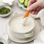 A hand dips a carrot stick into creamy ranch dressing, the perfect veggie dip. Sliced cucumbers, celery, and bowls of fresh herbs are arranged nearby on a light surface.