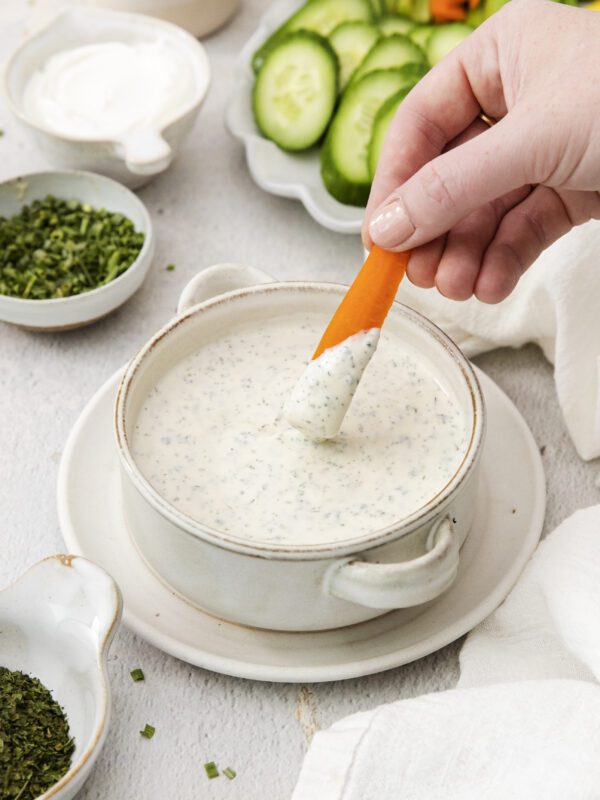 A hand dips a carrot stick into creamy ranch dressing, the perfect veggie dip. Sliced cucumbers, celery, and bowls of fresh herbs are arranged nearby on a light surface.