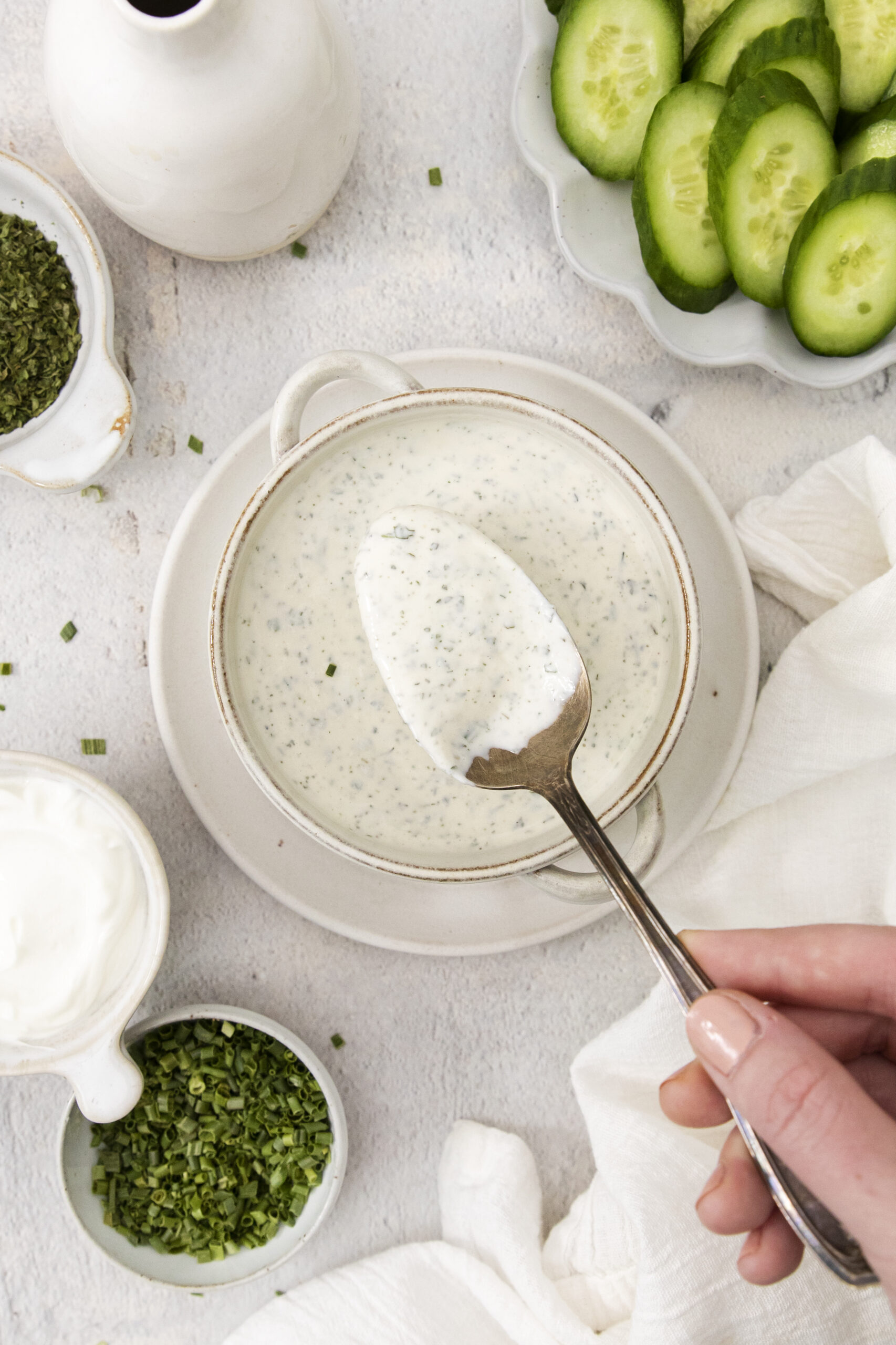 A hand holds a spoon above a bowl of creamy ranch dressing garnished with herbs, surrounded by fresh chopped chives, a plate of sliced cucumbers, and a bowl of sour cream—perfect for serving as a veggie dip on a light surface.