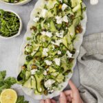 A person’s hands hold a platter of zucchini ribbon salad topped with feta cheese, pistachios, and greens. Nearby are bowls of feta, microgreens, fresh parsley, and half a lemon on a gray surface.