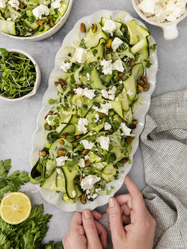A person’s hands hold a platter of zucchini ribbon salad topped with feta cheese, pistachios, and greens. Nearby are bowls of feta, microgreens, fresh parsley, and half a lemon on a gray surface.
