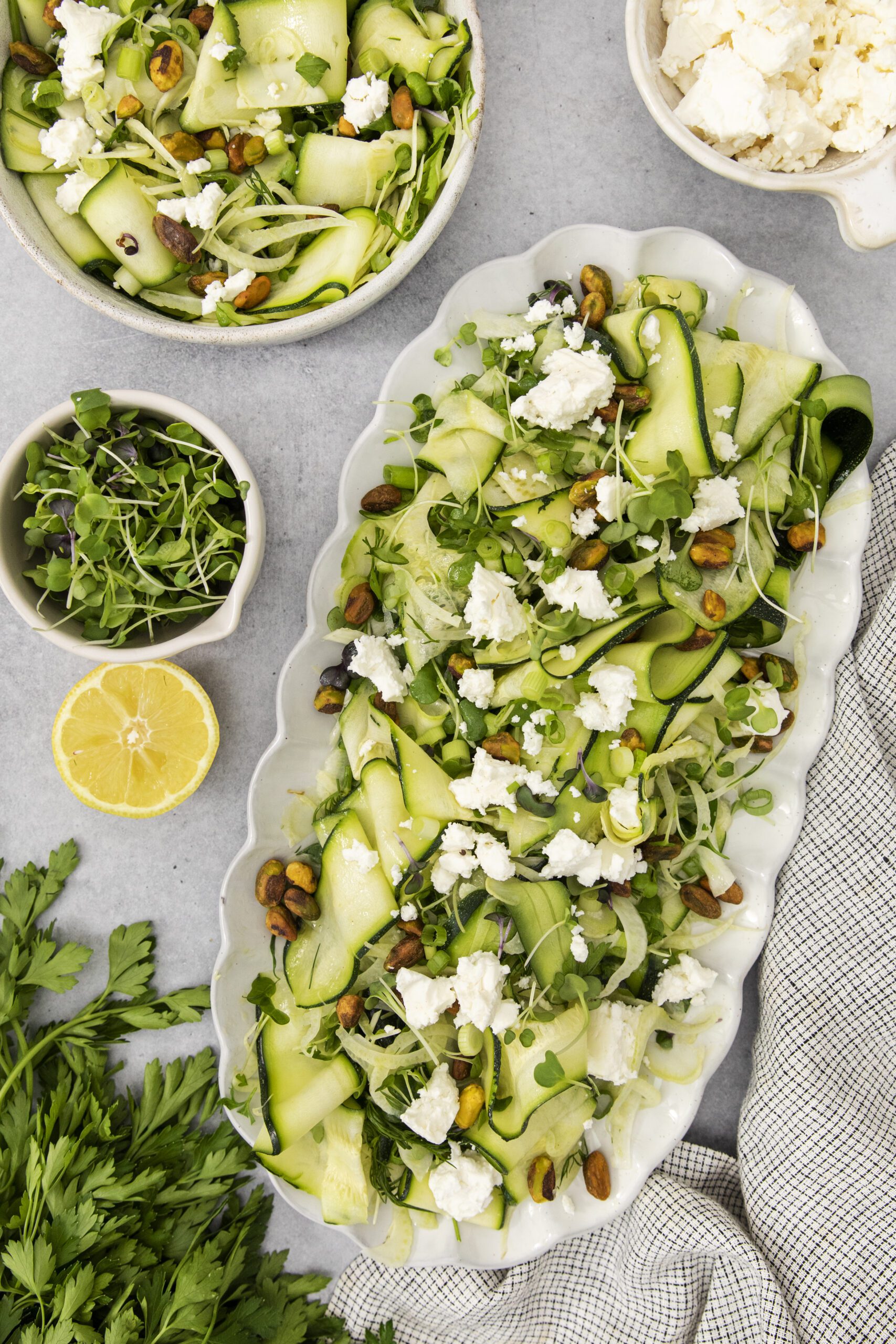 A platter of zucchini ribbon salad with crumbled cheese, pistachios, and microgreens, surrounded by a bowl of salad, a dish of cheese, microgreens, half a lemon, and fresh parsley on a light table.
