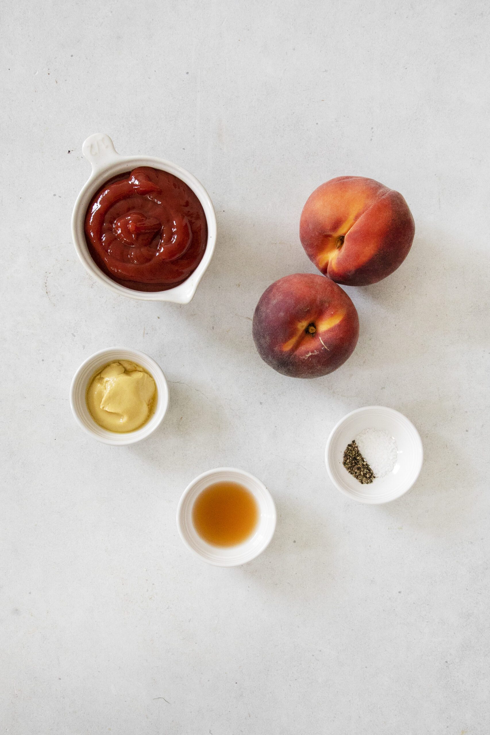 Five small bowls containing ketchup, mustard, vinegar, salt, and pepper are arranged on a light surface next to two whole peaches.