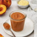 A glass jar filled with peach BBQ sauce sits on a white plate with a spoon beside it. Fresh peaches, a small bowl of mustard, and a striped cloth are nearby on a light surface.