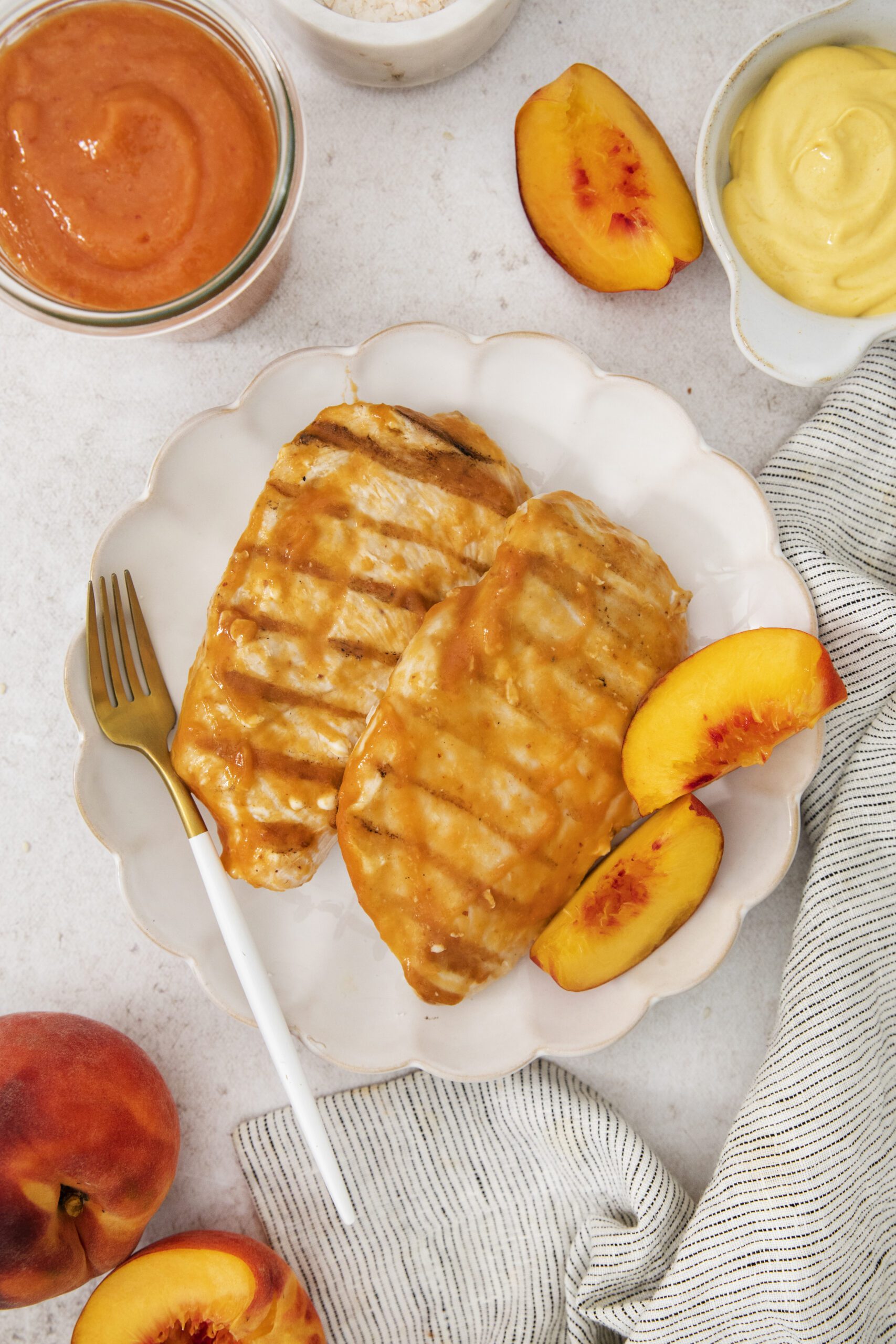 Grilled chicken breasts with peach glaze on a white plate, garnished with peach slices. A fork rests on the plate. Nearby are fresh peaches, a striped napkin, and small bowls of sauce.