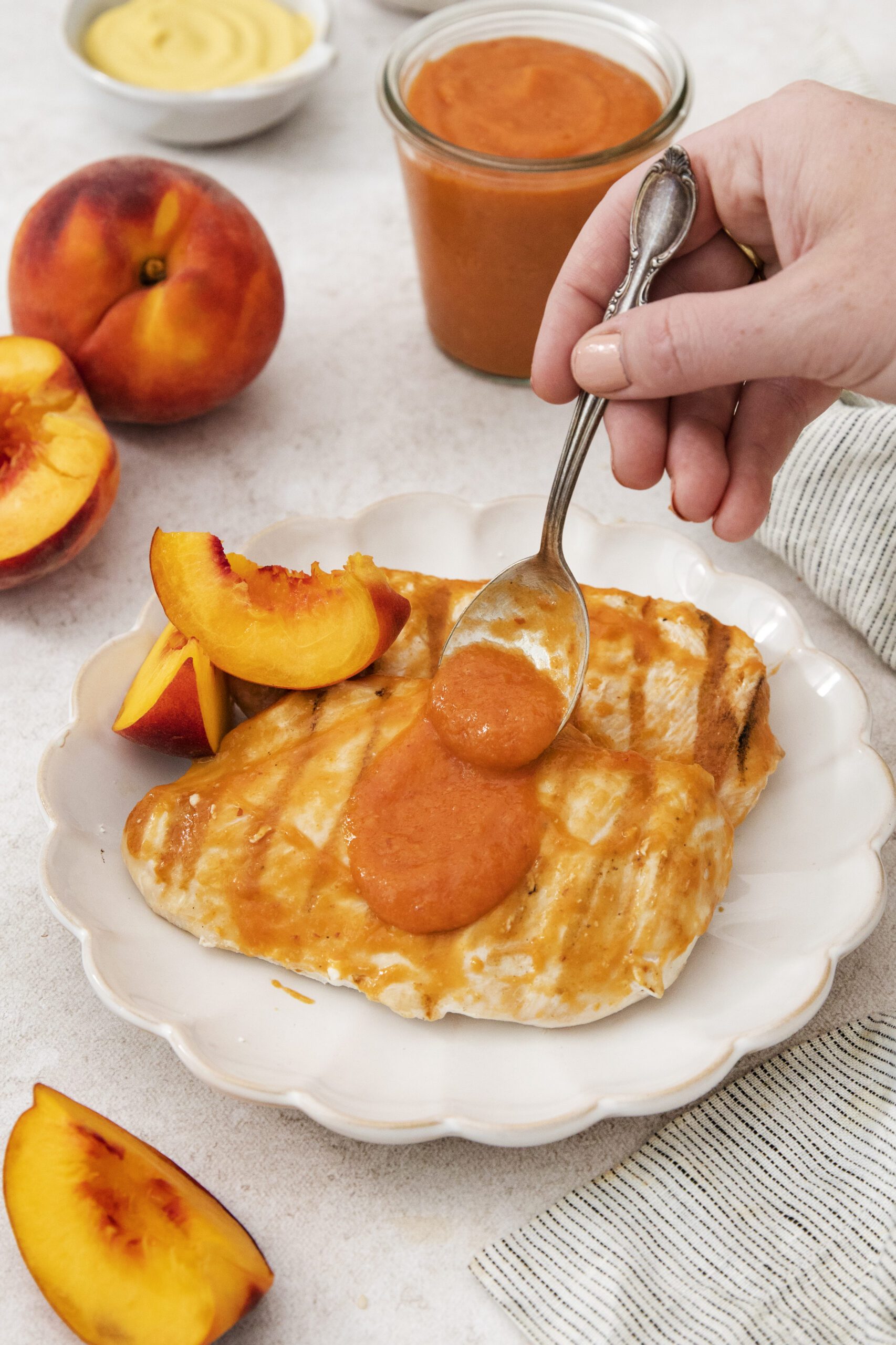 A hand spoons peach-colored sauce onto grilled chicken breast on a white plate, garnished with fresh peach slices. Whole and sliced peaches, a jar of sauce, and a striped napkin are nearby on the table.