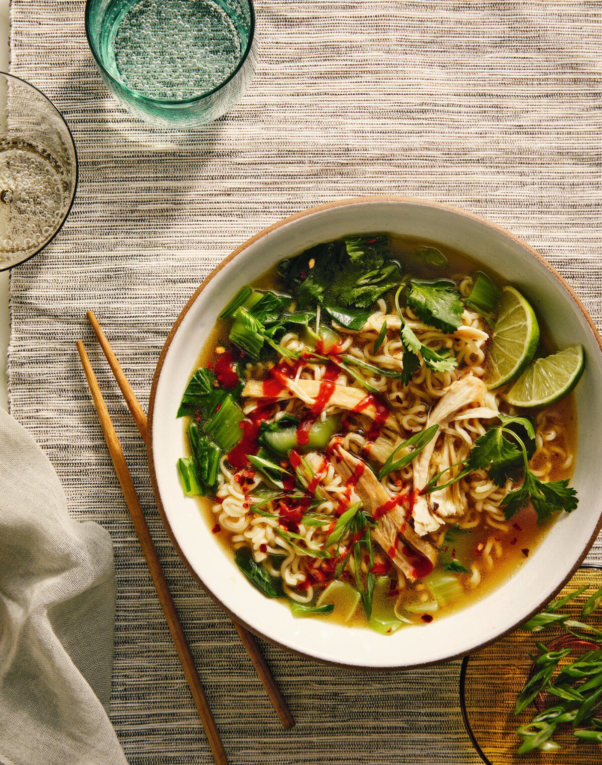 A bowl of ramen topped with shredded chicken, green onions, cilantro, lime wedges, and a drizzle of hot sauce sits on a striped cloth next to wooden chopsticks and glasses of sparkling water.