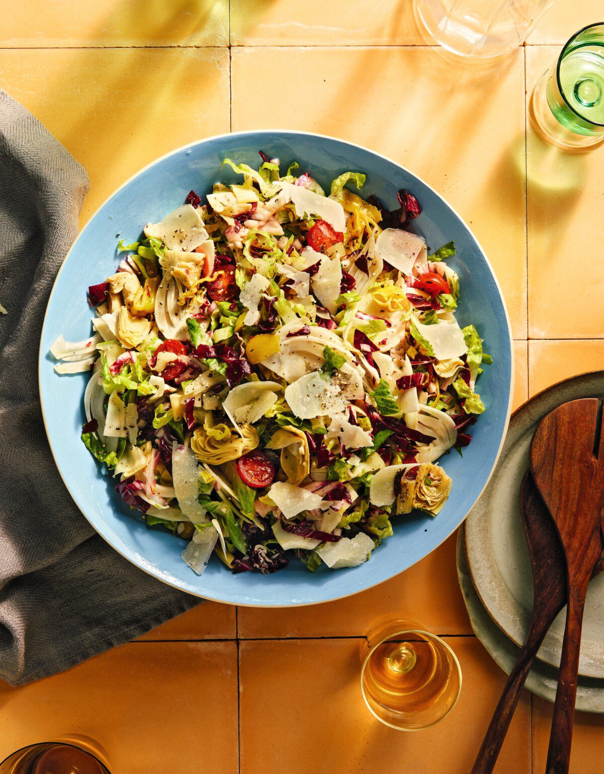 A blue bowl filled with a colorful salad of lettuce, cherry tomatoes, artichoke hearts, olives, radicchio, and shaved cheese, sitting on a yellow tiled table with glasses and wooden utensils nearby.