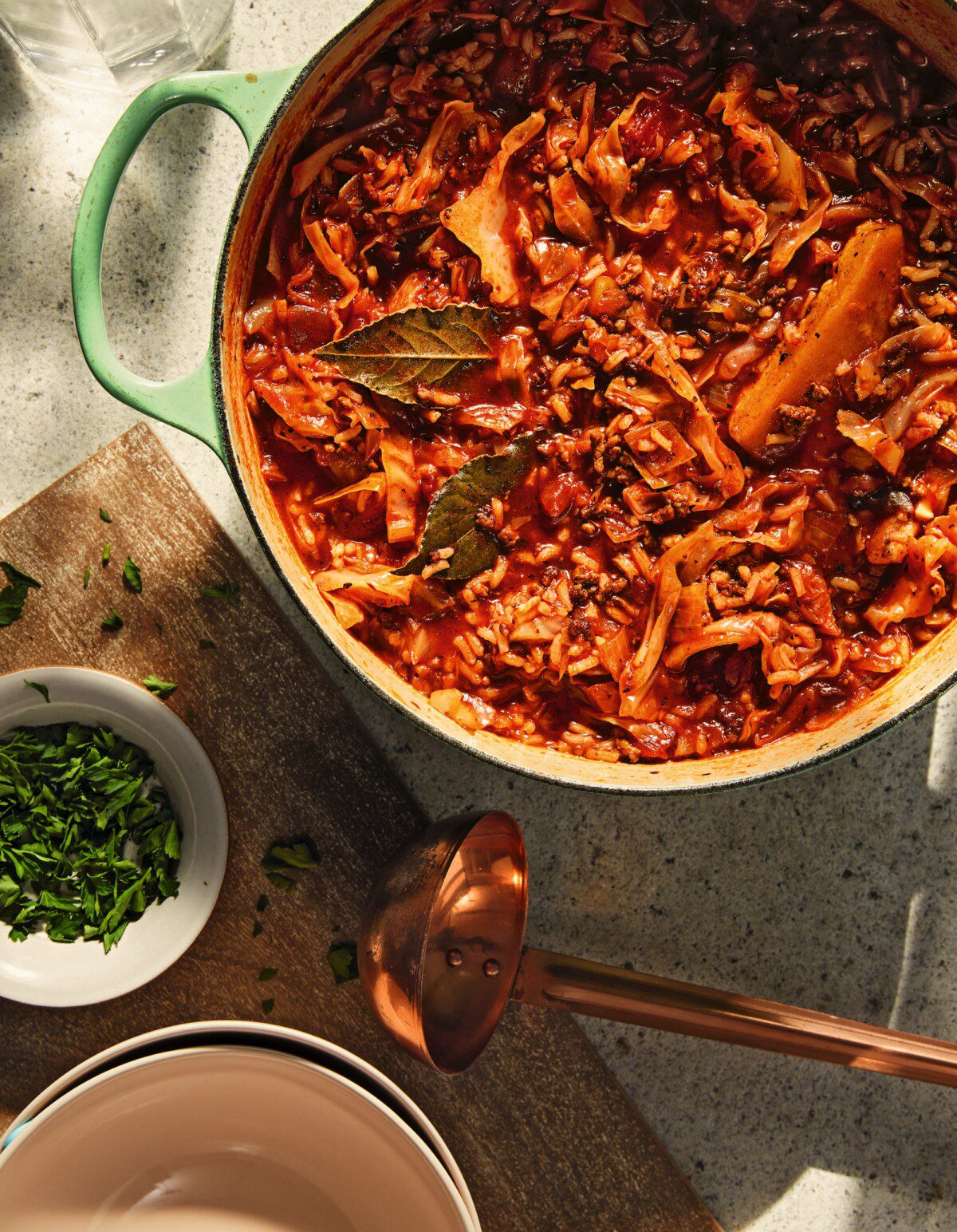 A pot of cabbage stew with bay leaves sits on a countertop beside a ladle, an empty bowl, and a small dish of chopped parsley. Sunlight highlights the rich red and green colors of the meal.