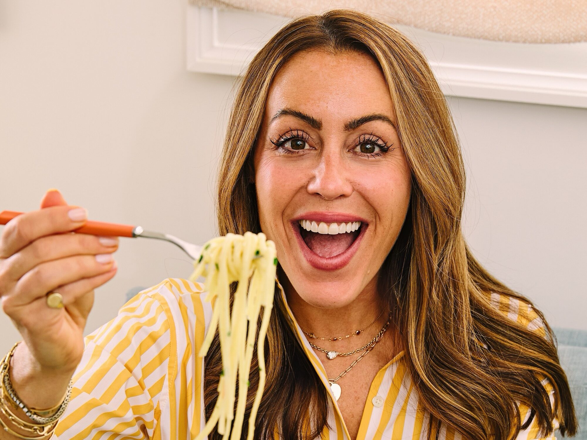 A woman in a yellow striped shirt sits on a couch, smiling and holding a forkful of spaghetti above a green plate of pasta with pieces of salmon.