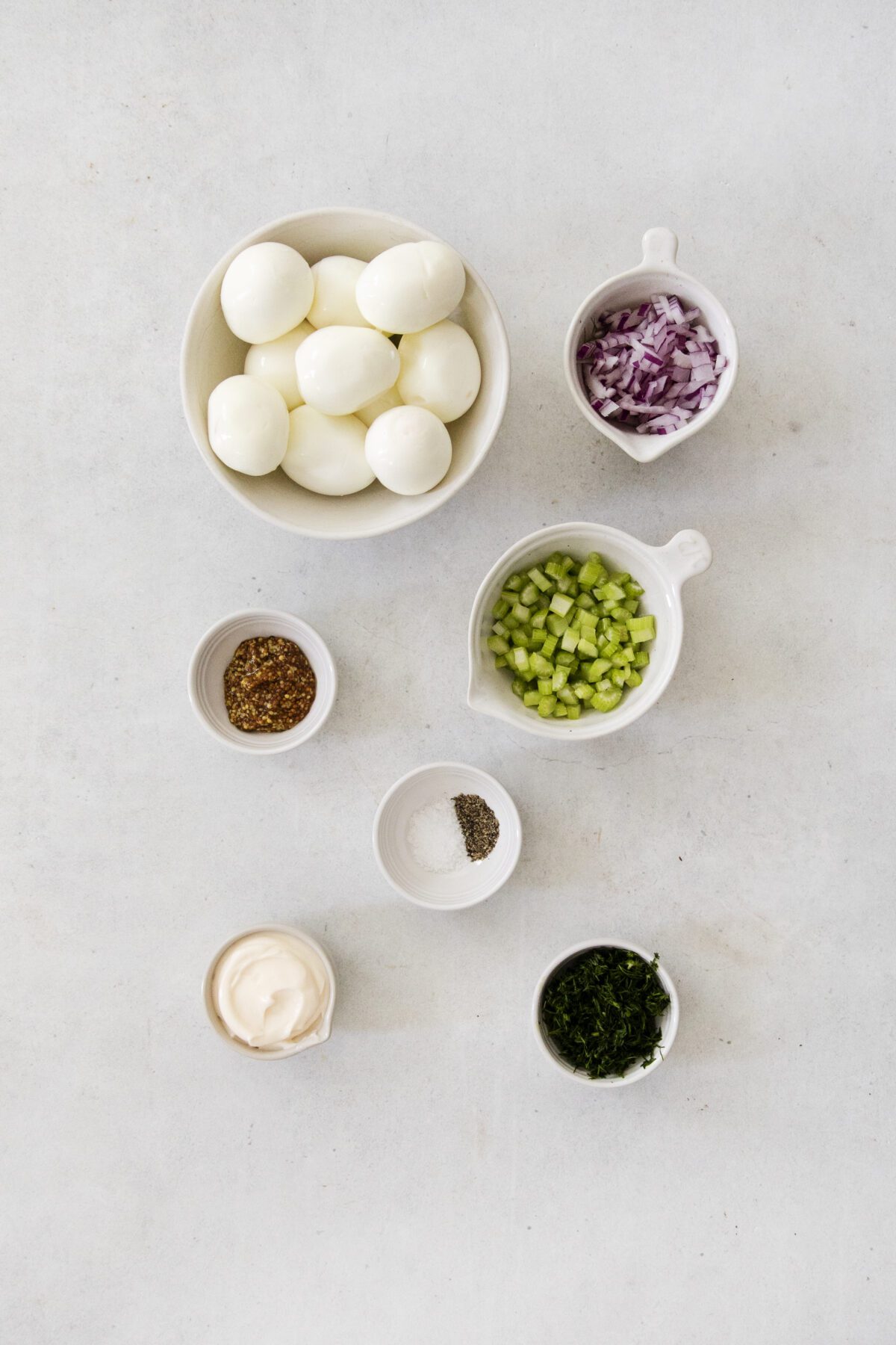 A flat lay of small bowls containing hard-boiled eggs, chopped red onion, diced celery, chopped herbs, mayonnaise, seasoning, and salt and pepper on a light surface.