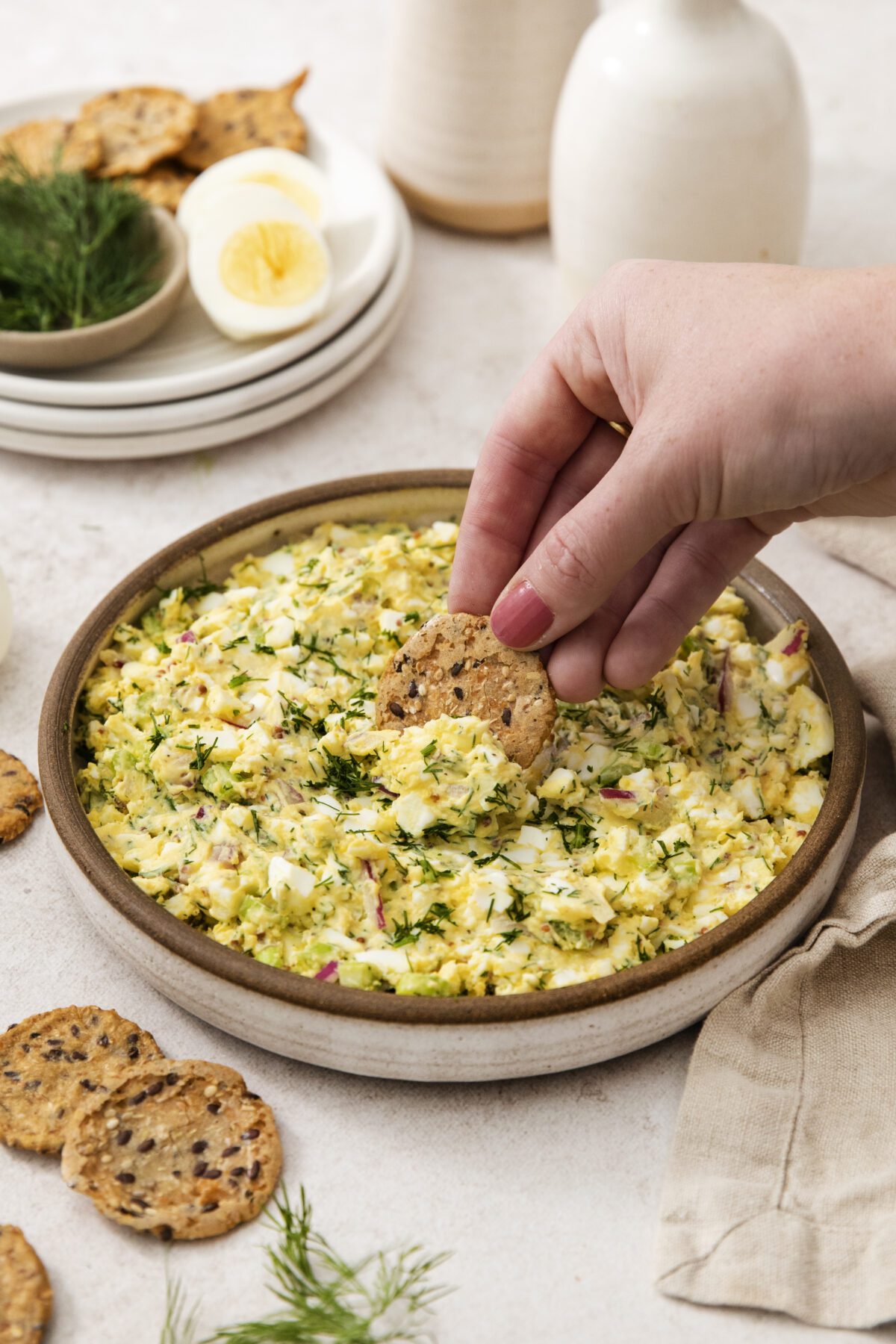 A hand dips a seeded cracker into a bowl of creamy egg salad garnished with fresh dill. Sliced hard-boiled eggs and more crackers are arranged on plates in the background.