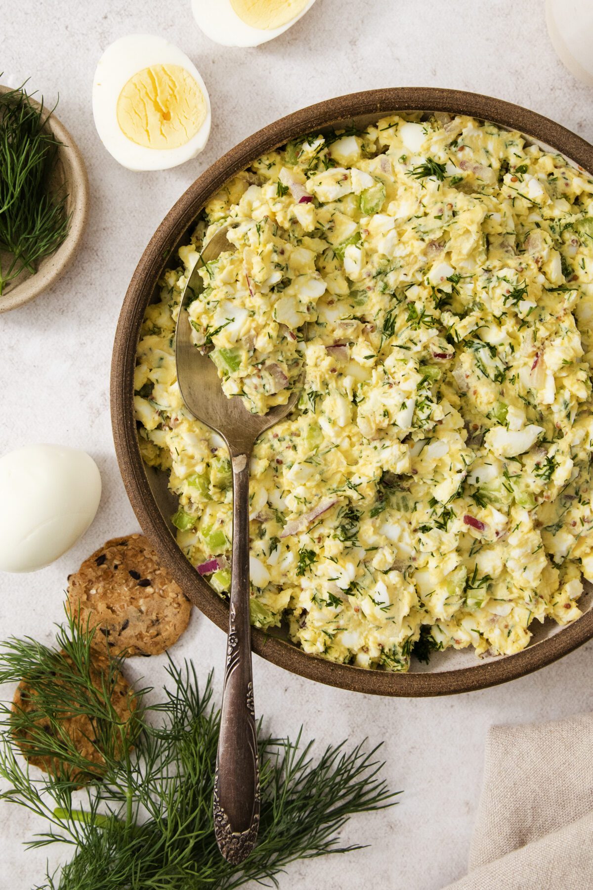 A bowl of egg salad with chopped dill and red onion, served with a spoon. Surrounding the bowl are hard-boiled eggs, crackers, and fresh dill on a light surface.