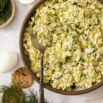 A bowl of egg salad with chopped dill and red onion, served with a spoon. Surrounding the bowl are hard-boiled eggs, crackers, and fresh dill on a light surface.