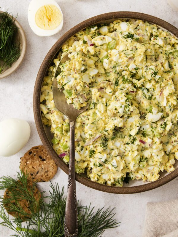 A bowl of egg salad with chopped dill and red onion, served with a spoon. Surrounding the bowl are hard-boiled eggs, crackers, and fresh dill on a light surface.