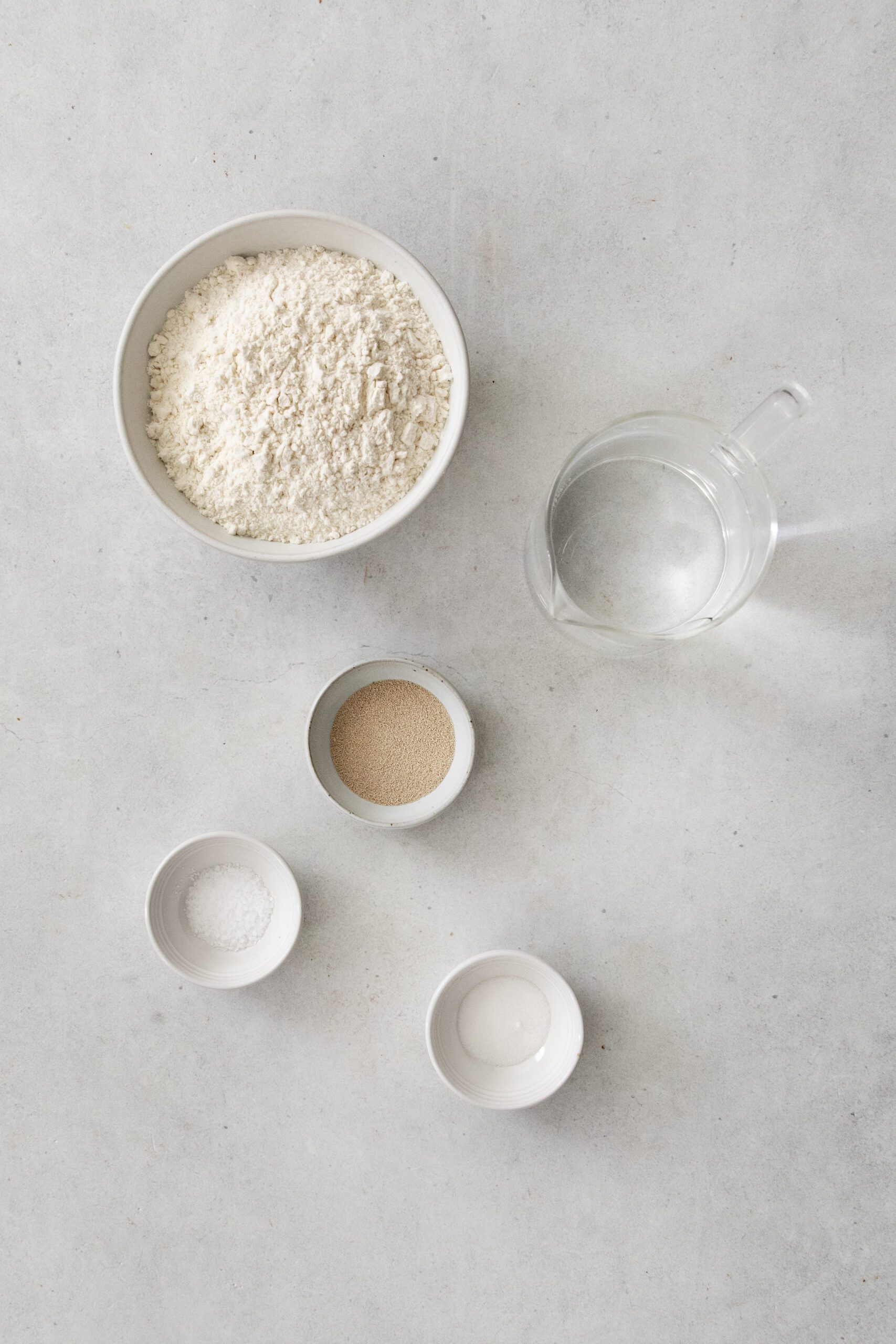 A bowl of flour, a glass jug of water, a small bowl of active dry yeast, a small bowl of salt, and a small bowl of sugar arranged on a light gray surface.