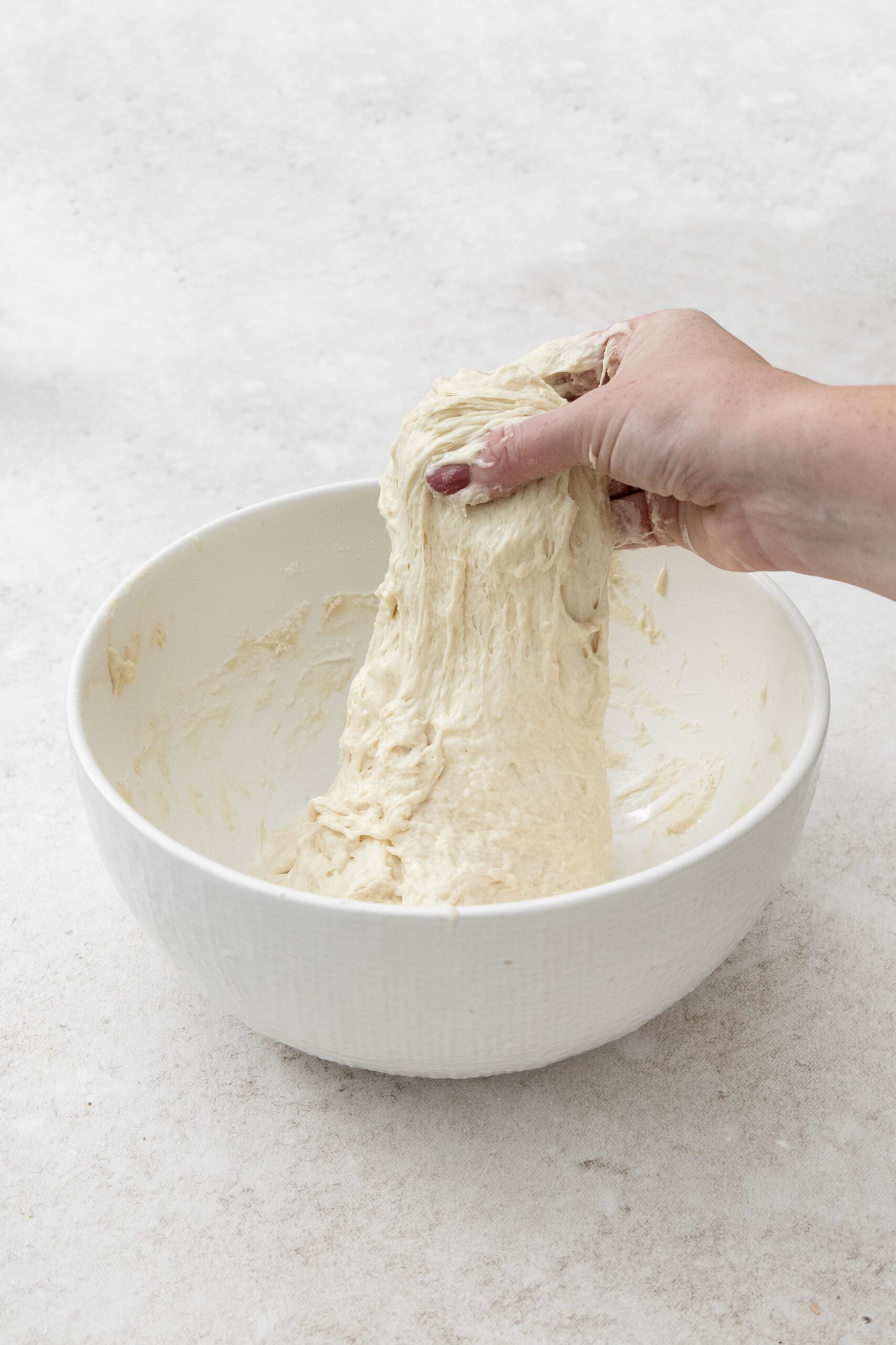 A hand stretches sticky bread dough upwards from a white mixing bowl on a light-colored surface.