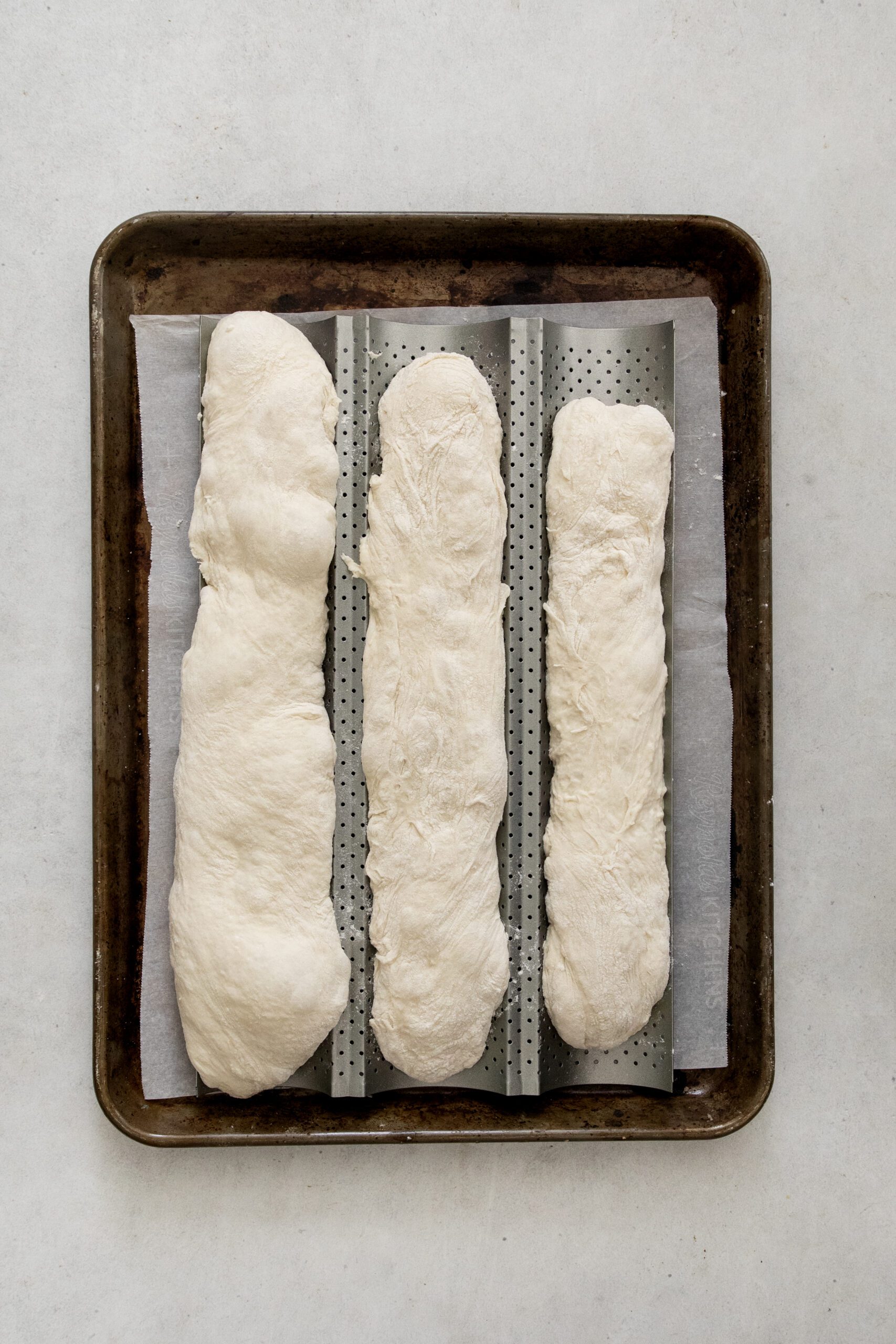 Three unbaked baguette loaves made from dough rest on a perforated baking tray lined with parchment paper, ready to be baked.