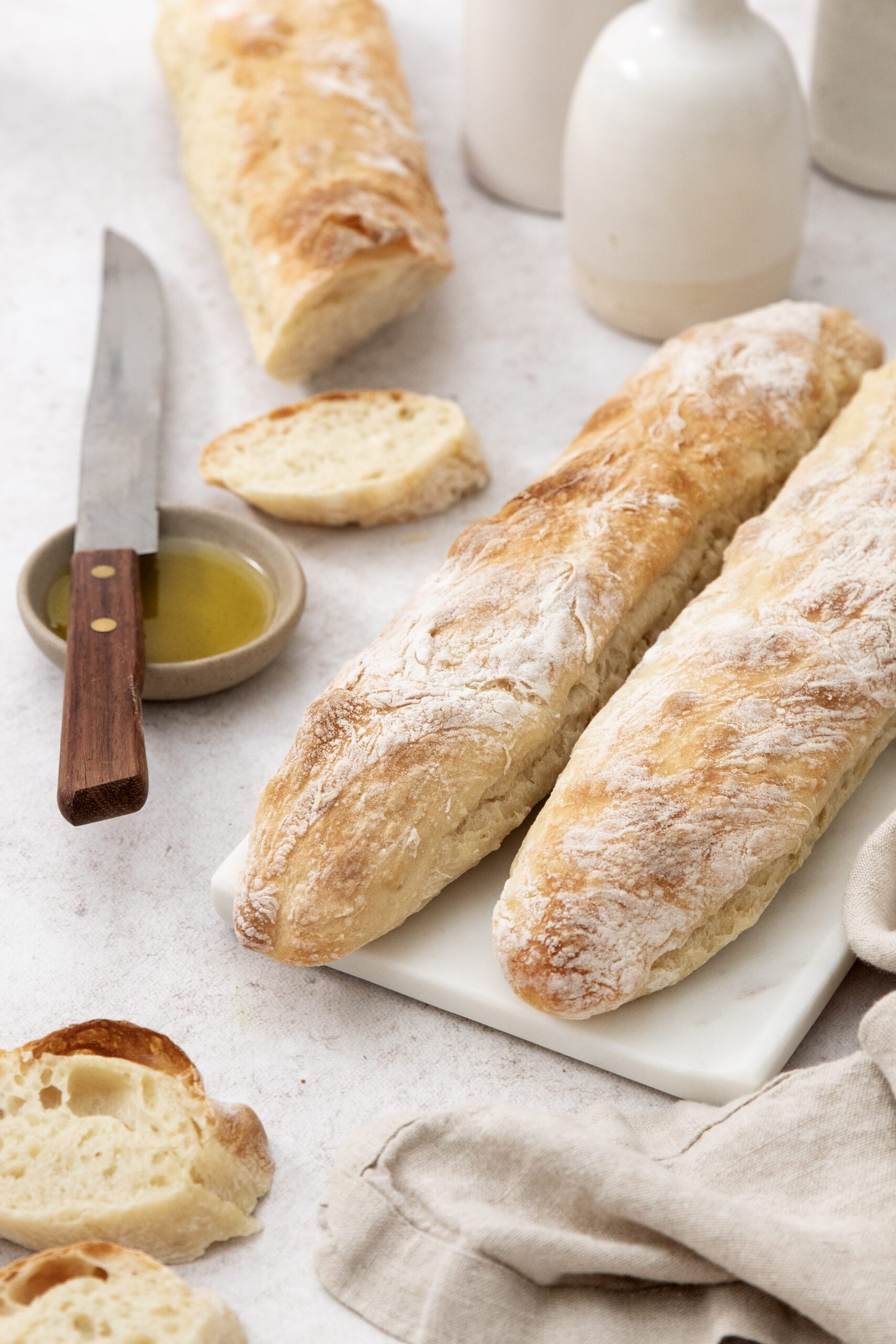 Two rustic baguettes rest on a white platter with slices nearby. A knife, a small dish of olive oil, and cream-colored ceramics are on the table, along with a beige cloth. The scene is light and inviting.
