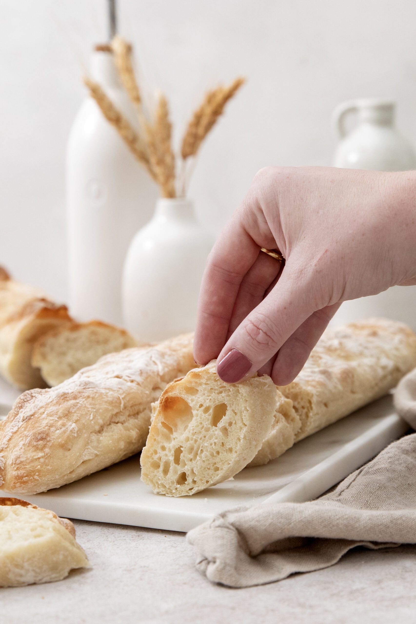 A hand with painted nails holds a slice of rustic bread above a platter with more bread loaves. White ceramic vases and a jug are in the background, with wheat stalks displayed in one vase.