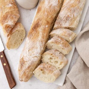 A rustic baguette is partially sliced on a white marble board, accompanied by a knife, a beige napkin, a small bowl of olive oil, and a vase on a light surface.