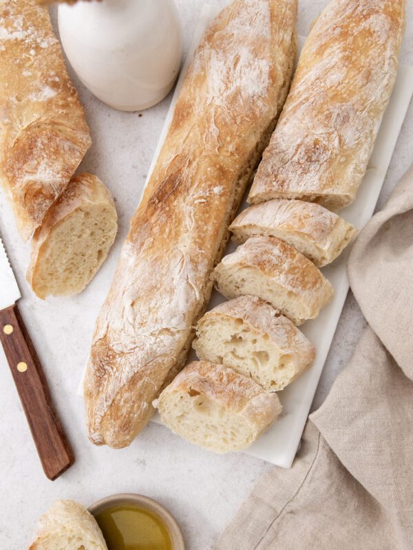 A rustic baguette is partially sliced on a white marble board, accompanied by a knife, a beige napkin, a small bowl of olive oil, and a vase on a light surface.