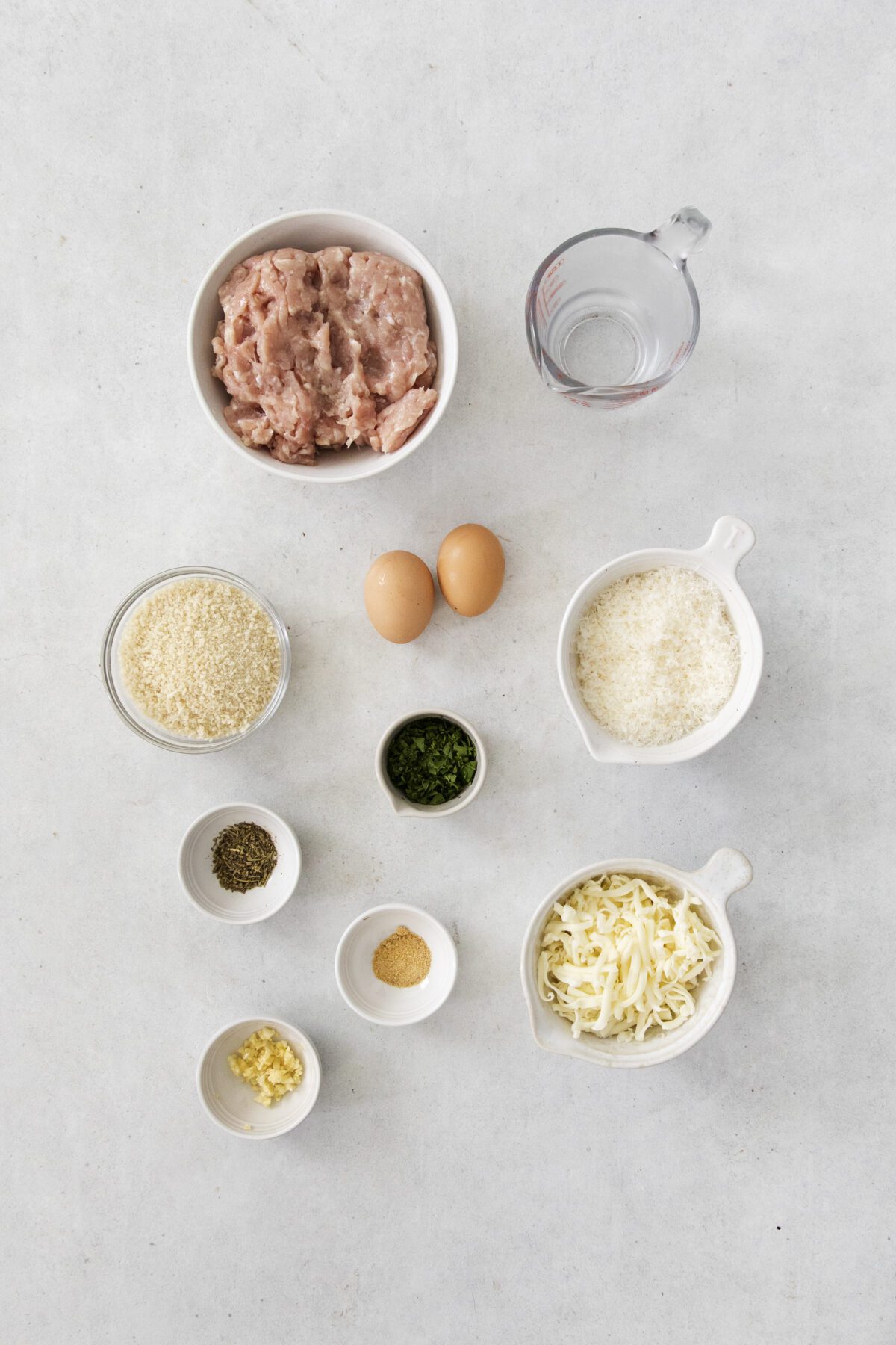 A flat lay of meatball ingredients: ground meat, water in a measuring cup, two eggs, breadcrumbs, grated cheese, chopped herbs, shredded cheese, dried herbs, garlic, and a small pile of seasoning on a light surface.