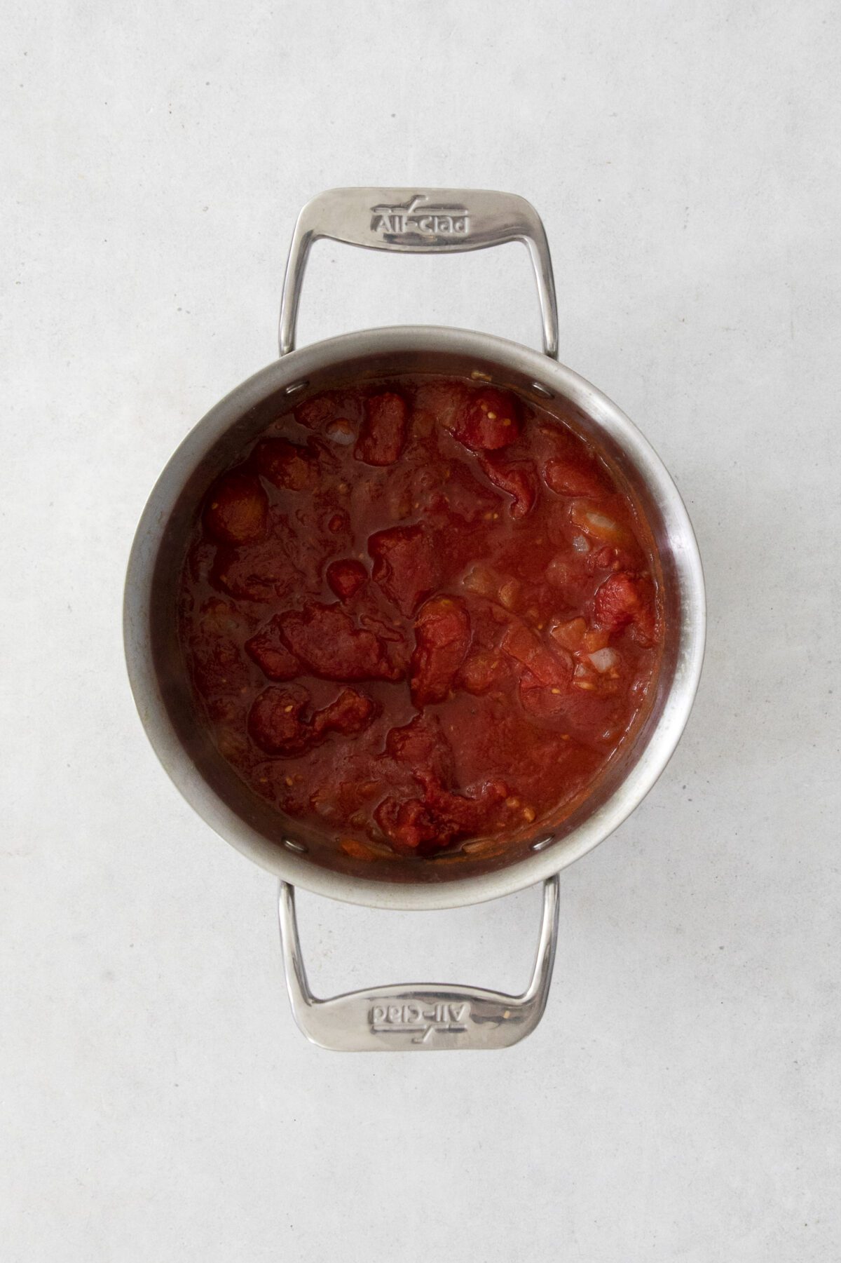 A stainless steel pot filled with chunky red tomato sauce, visible tomato pieces, and onions, placed on a light gray surface.