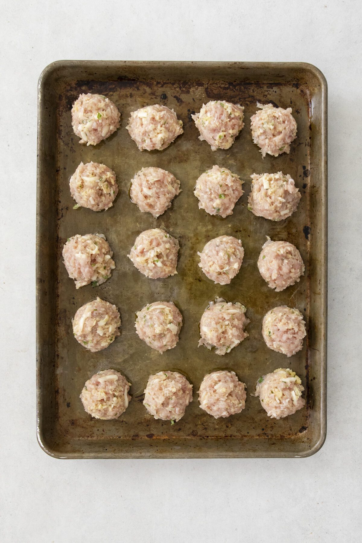 A baking tray holds 20 evenly spaced raw meatballs made from a mixture with visible diced vegetables, ready to be cooked. The tray sits on a light-colored surface.