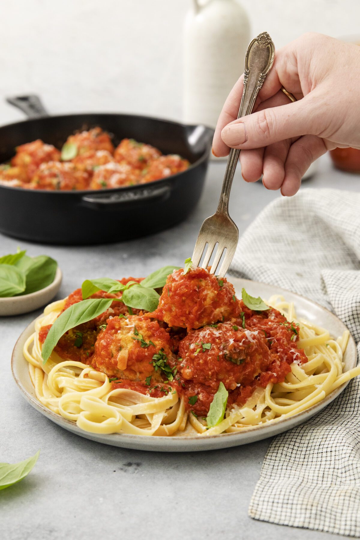 A hand holding a fork is about to pick up a meatball from a plate of pasta topped with tomato sauce and fresh basil. A skillet of more meatballs is in the background. A cloth napkin is beside the plate.