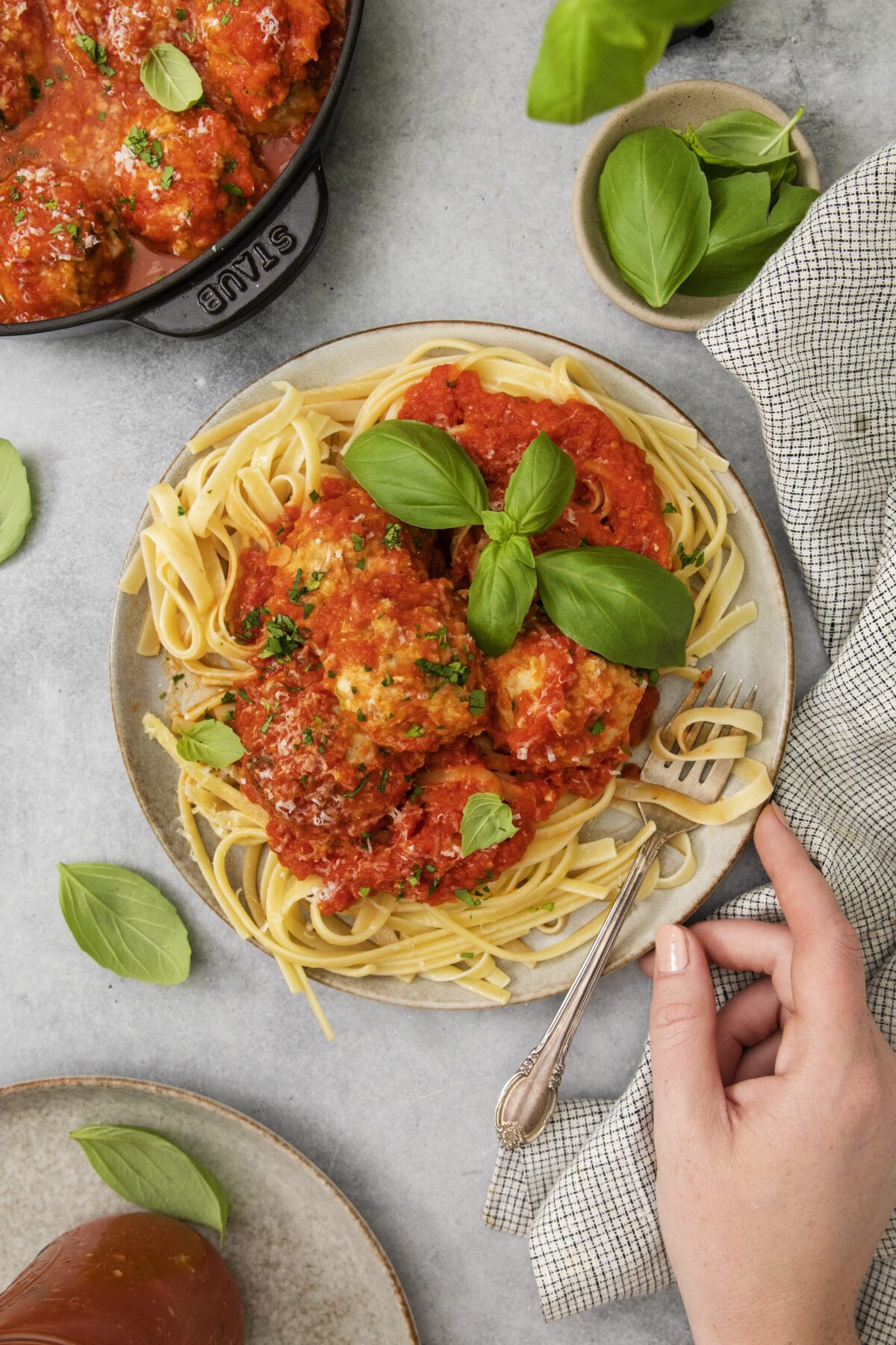 A plate of pasta topped with meatballs in tomato sauce, garnished with fresh basil, sits on a table. A hand holds a fork, and a pot of meatballs, basil leaves, and a napkin are nearby.