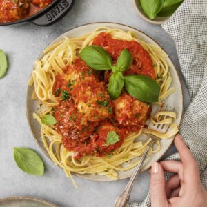 A plate of pasta topped with meatballs in tomato sauce, garnished with fresh basil, sits on a table. A hand holds a fork, and a pot of meatballs, basil leaves, and a napkin are nearby.