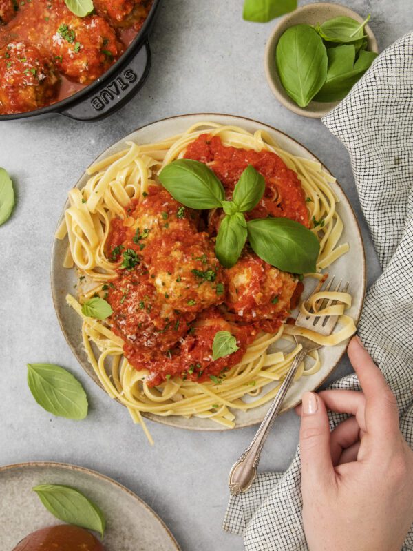 A plate of pasta topped with meatballs in tomato sauce, garnished with fresh basil, sits on a table. A hand holds a fork, and a pot of meatballs, basil leaves, and a napkin are nearby.