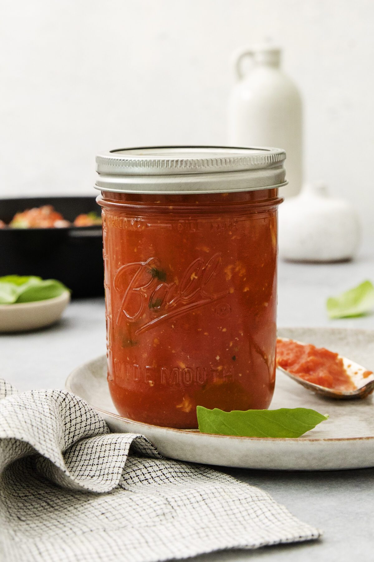 A glass jar filled with tomato sauce sits on a plate next to a spoon with sauce, a bay leaf, and a checkered cloth. Blurred basil leaves and containers are in the background.