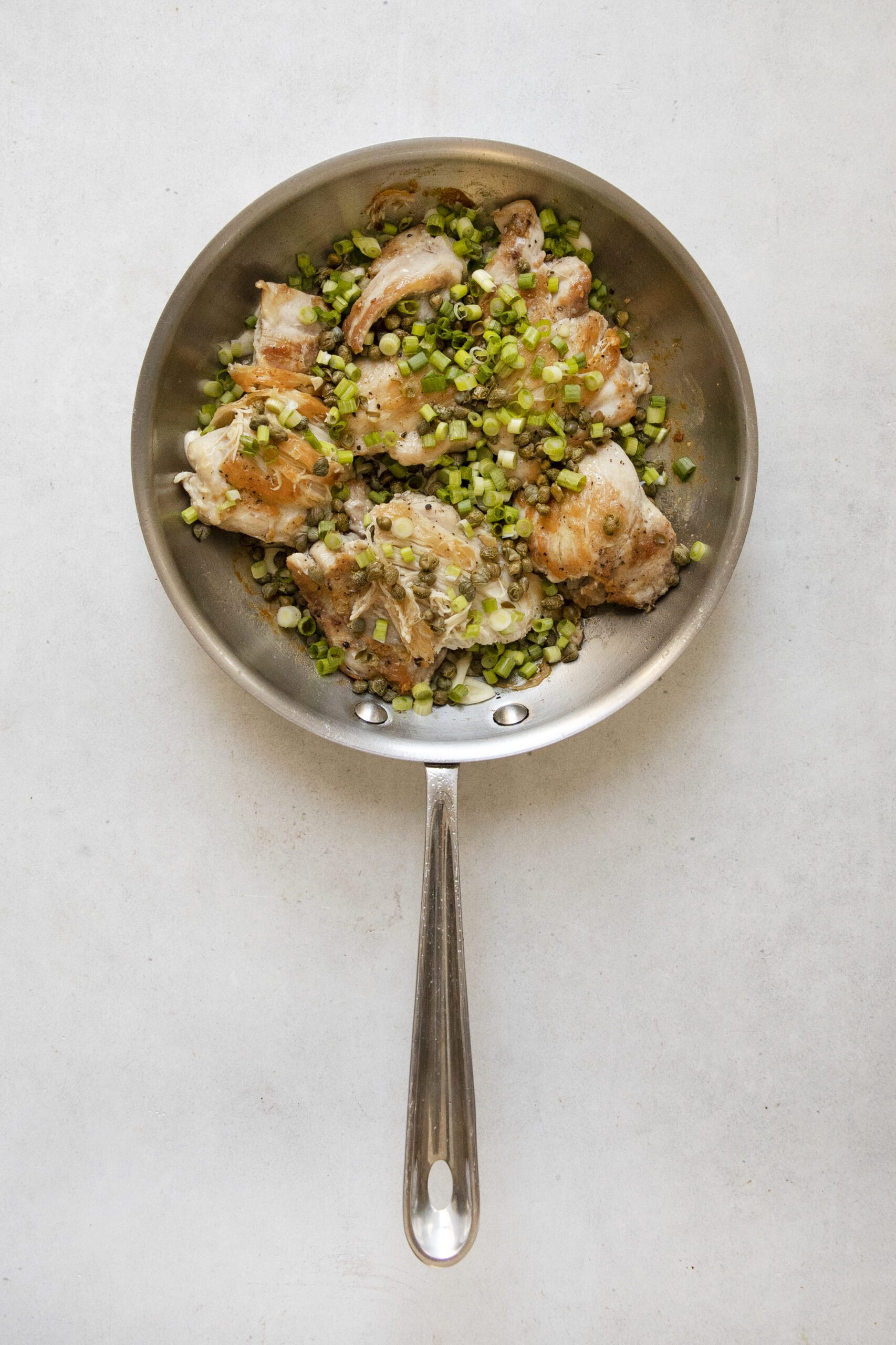 A stainless steel skillet with cooked chicken pieces topped with chopped green onions, set on a light surface.