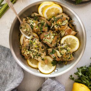 A pan of cooked chicken garnished with fresh herbs, capers, and lemon slices. A spoon rests inside the pan. Sliced lemons, parsley, and a striped cloth napkin are arranged around the pan on a light surface.