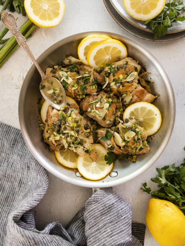 A pan of cooked chicken garnished with fresh herbs, capers, and lemon slices. A spoon rests inside the pan. Sliced lemons, parsley, and a striped cloth napkin are arranged around the pan on a light surface.