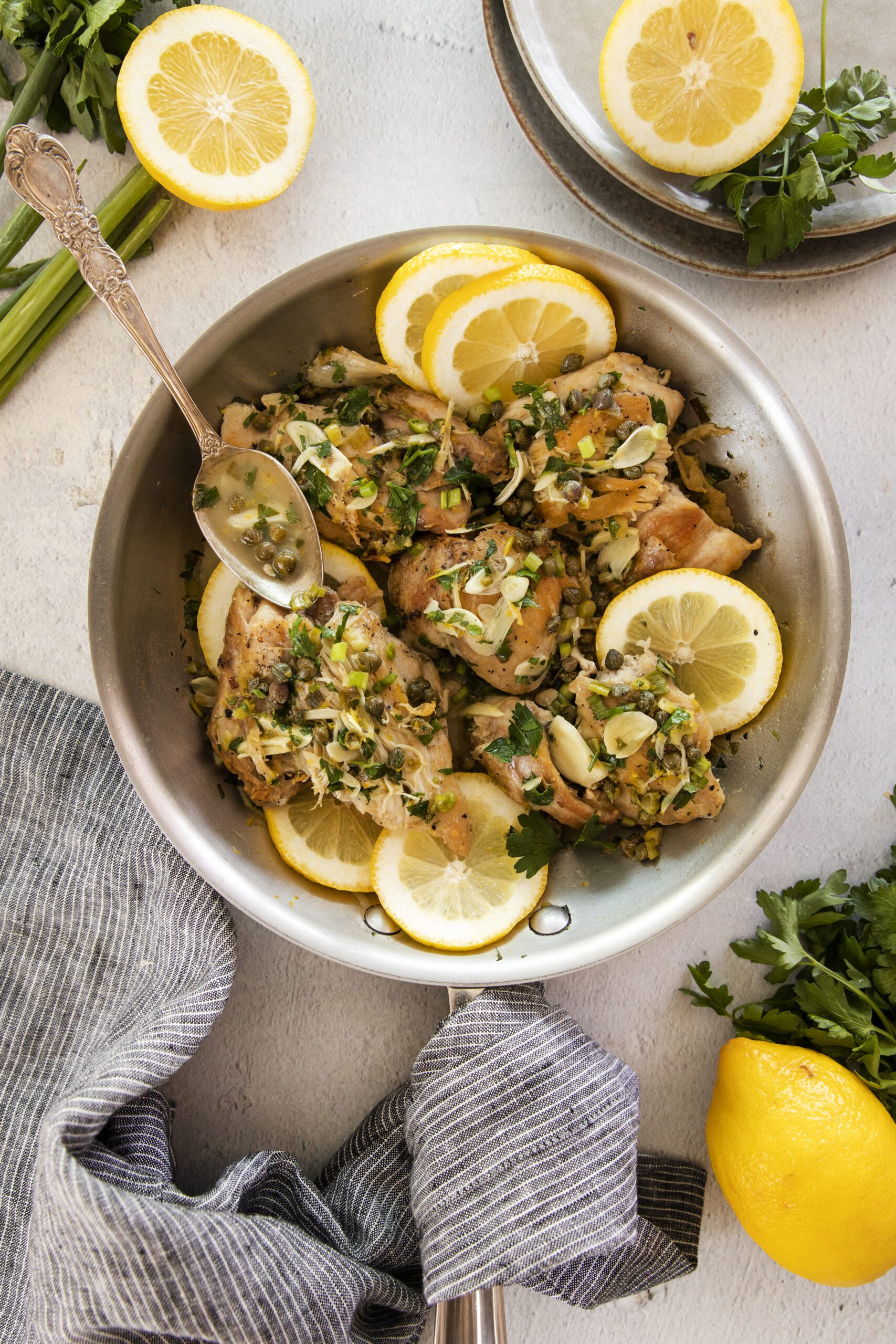 A skillet filled with cooked chicken topped with chopped herbs, garlic, and capers, garnished with lemon slices. A spoon rests in the pan, with fresh parsley, lemon, and a striped towel nearby.
