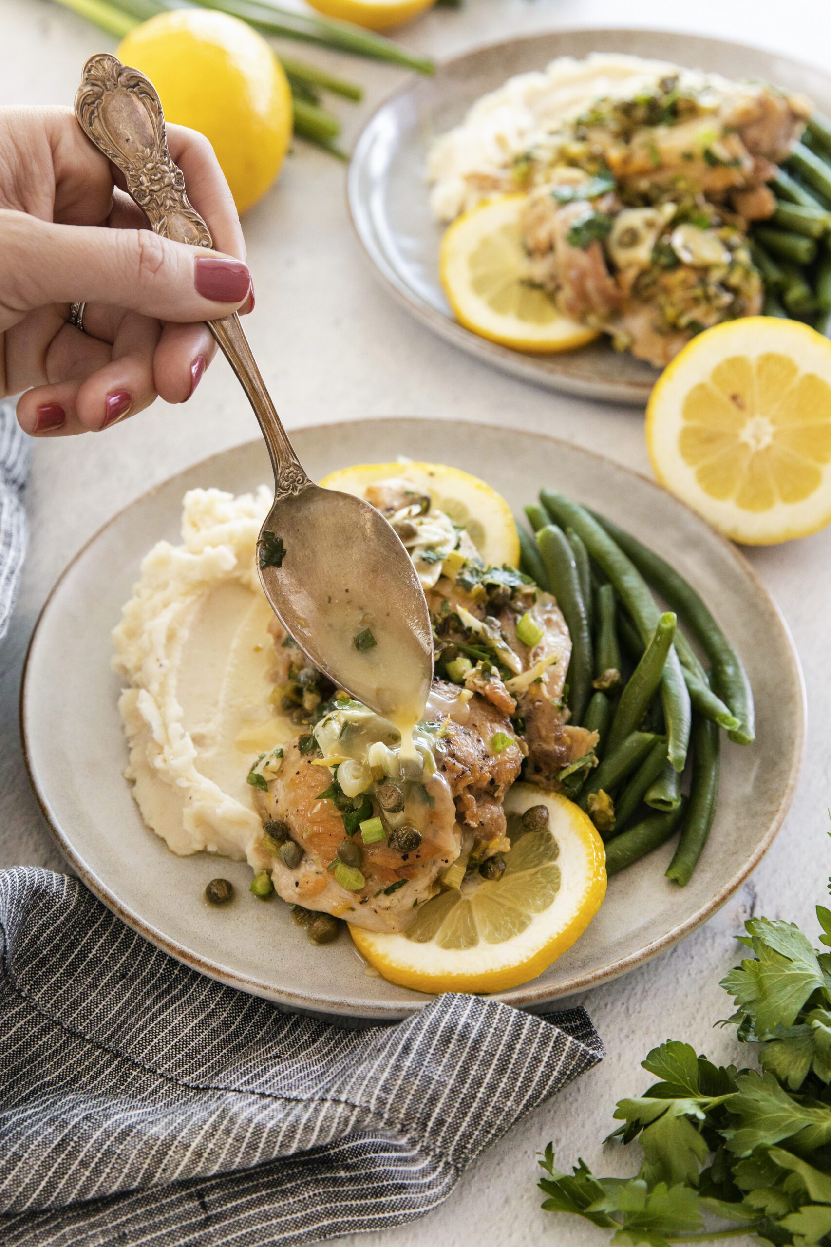 A hand holds a spoon over a plate of chicken piccata with sauce, mashed potatoes, green beans, and lemon slices. Another similar plate is in the background, along with fresh lemons and parsley on the table.