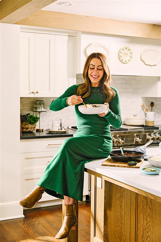 A woman in a long green dress and brown boots sits on a kitchen counter, smiling as she eats from a bowl. The kitchen has white cabinets, wooden accents, and various bowls and utensils on the counter.