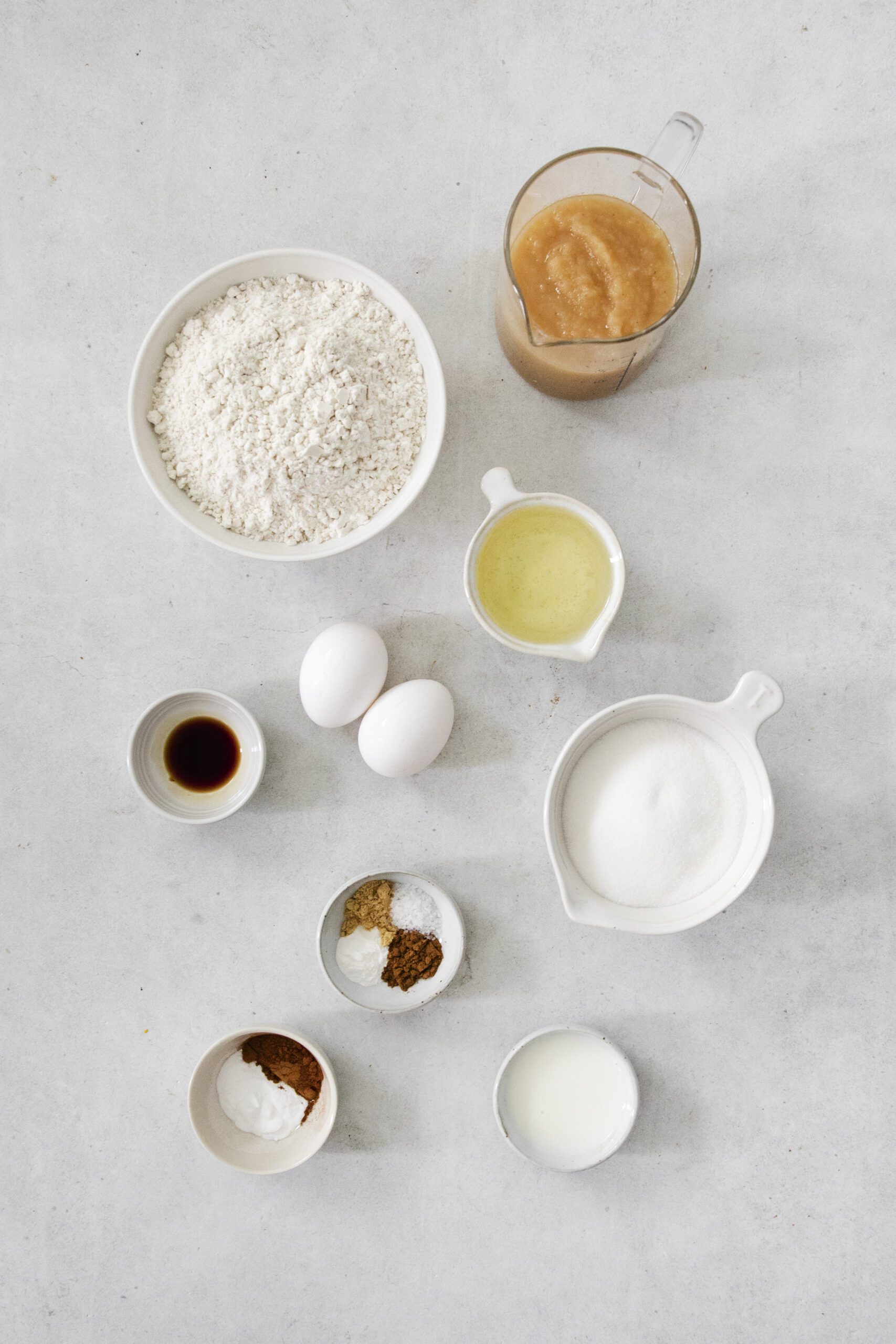 A flat lay of baking ingredients on a light surface, including flour, applesauce, two eggs, egg whites, sugar, milk, vanilla extract, baking powder, baking soda, salt, and spices in separate bowls and cups.