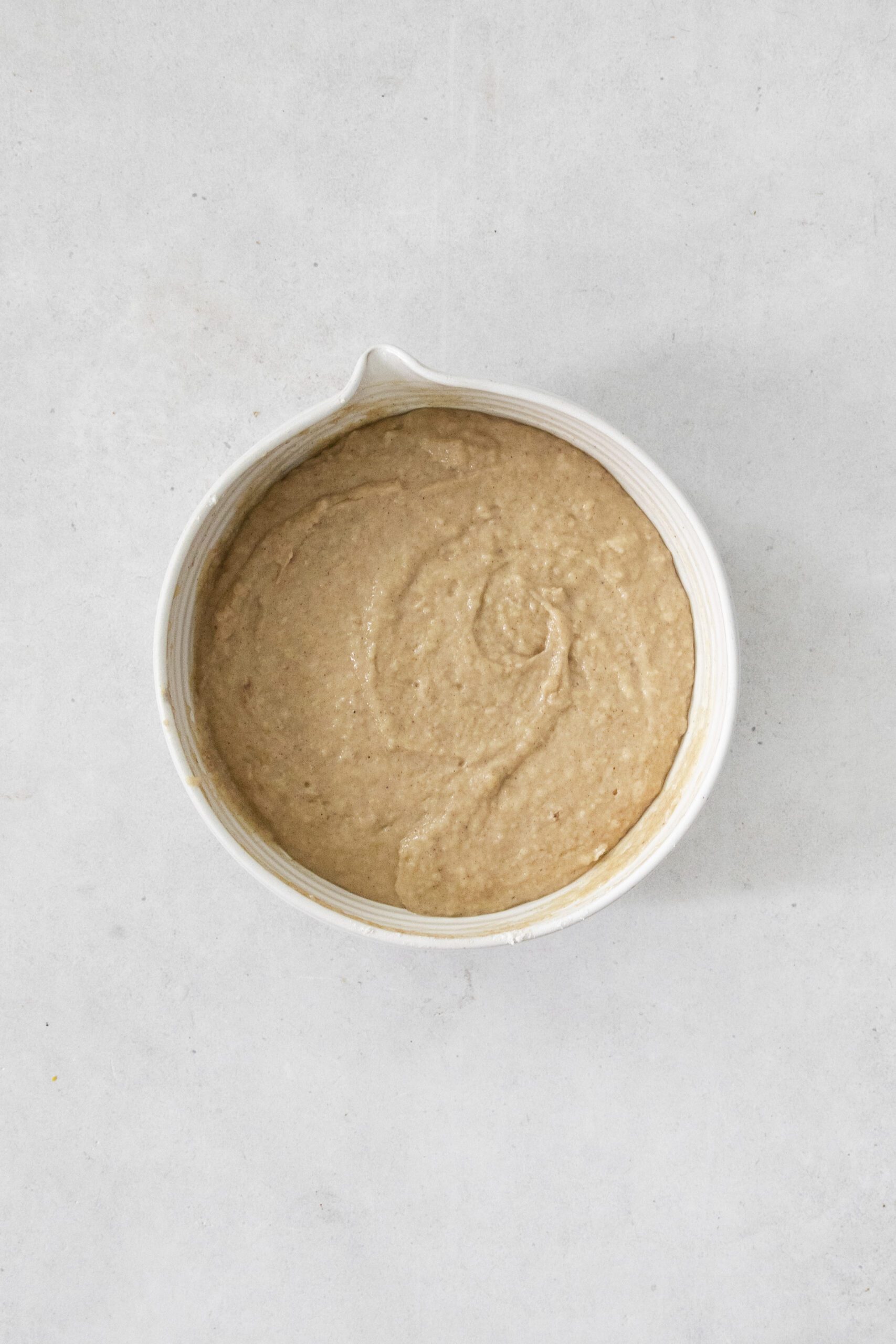 A white mixing bowl filled with smooth, light brown batter sits on a light gray surface.